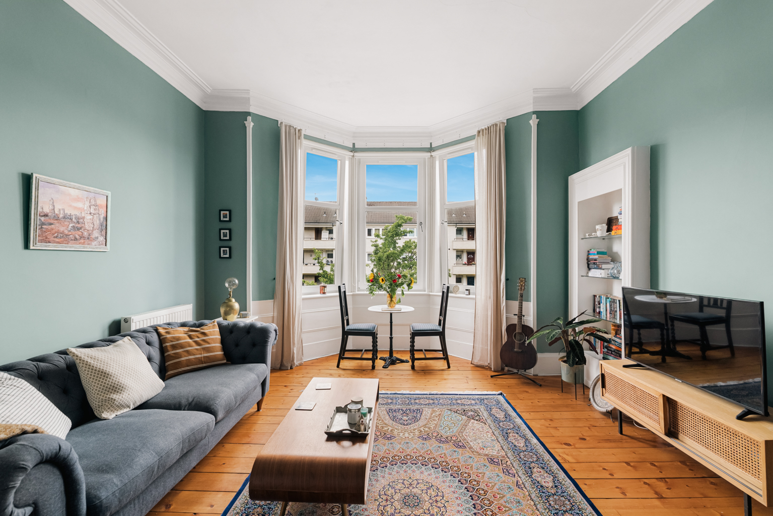 Bright, well-presented tenement flat lounge on Thornwood Gardens in Glasgow's West End, with sage-green walls, original cornicing, a bay window with bistro table and fresh flowers, and stripped pine floorboards