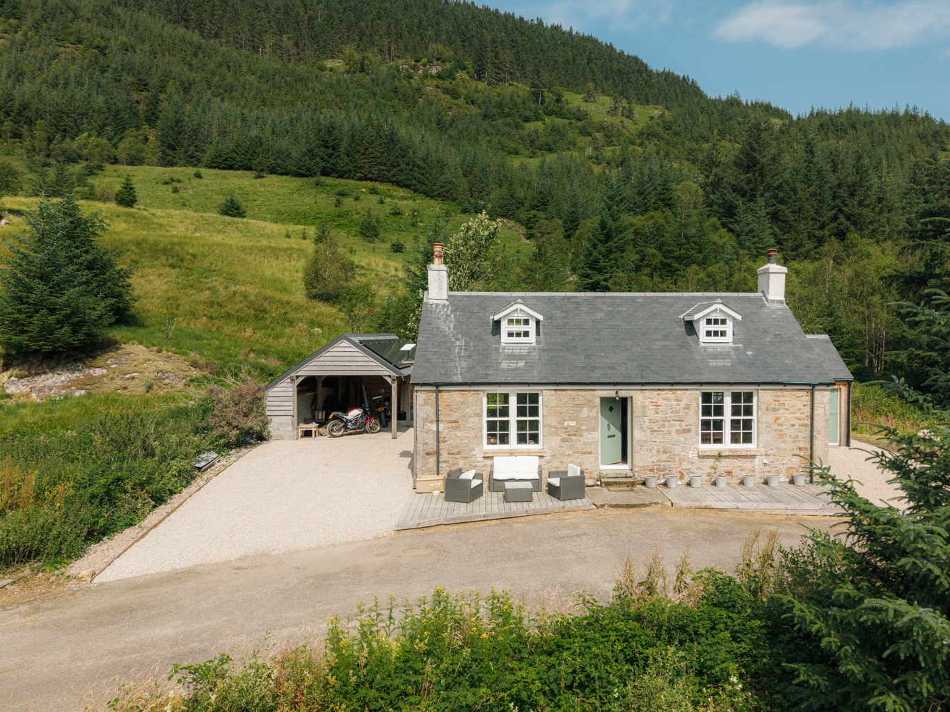 Traditional stone cottage for sale near Glenfinnan, Polloch, with a motorbike parked in the carport and forested hills rising behind