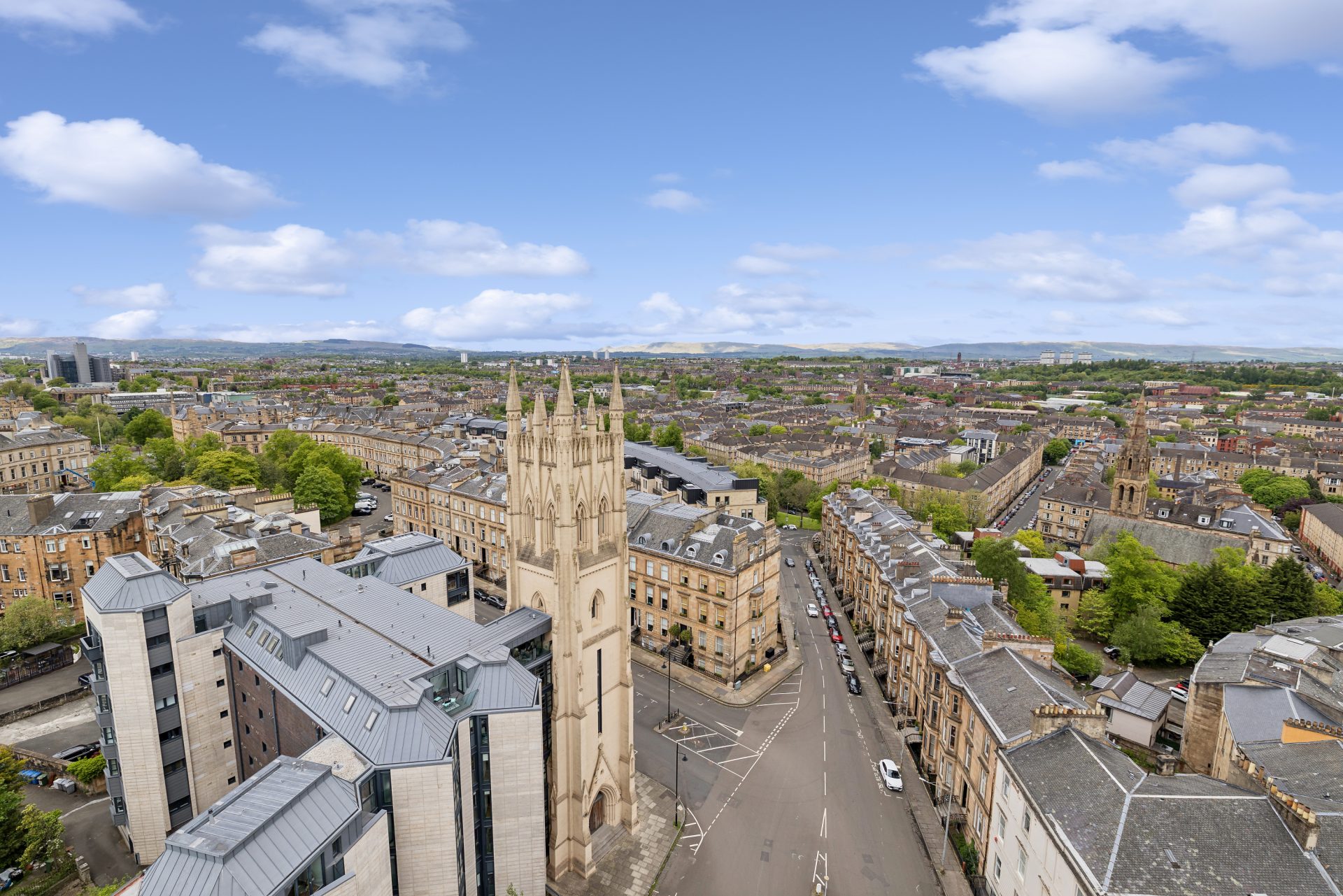 Aerial view over Glasgow's West End, with blond sandstone terraces, a Gothic church tower and a modern apartment block, and hills on the horizon